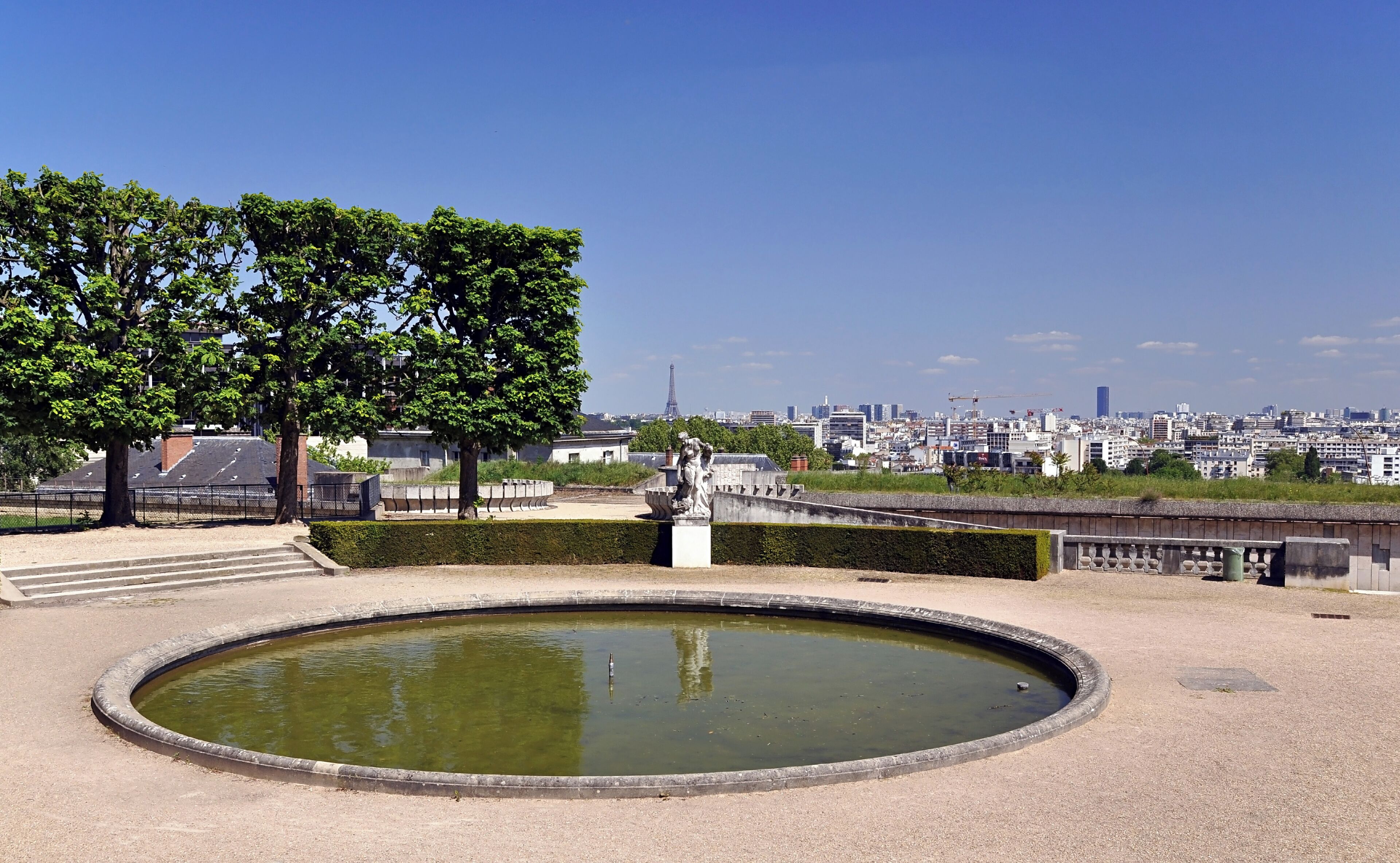 A park with a circular pond, trees, and a statue. In the background, there is a cityscape with buildings and the Eiffel Tower.