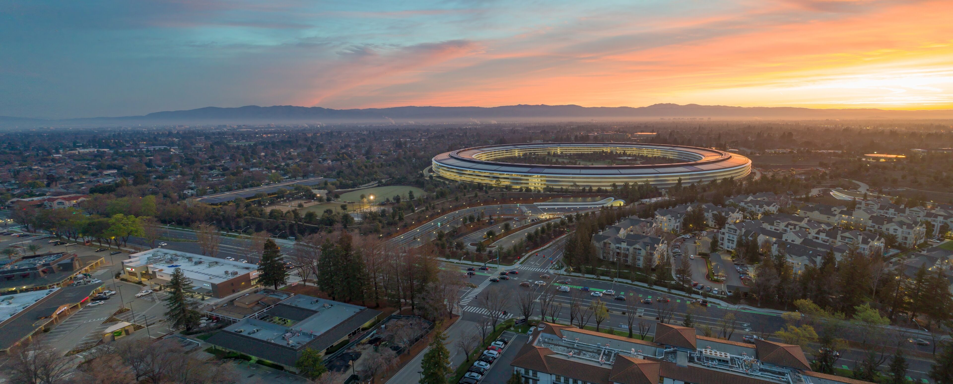 Aerial view of Apple Park headquarters at sunrise in Cupertino, California, USA. The photo captures the iconic circular building and surrounding landscape.