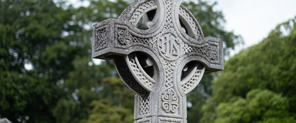 A detailed stone Celtic cross with intricate carvings, set against a backdrop of lush green trees.