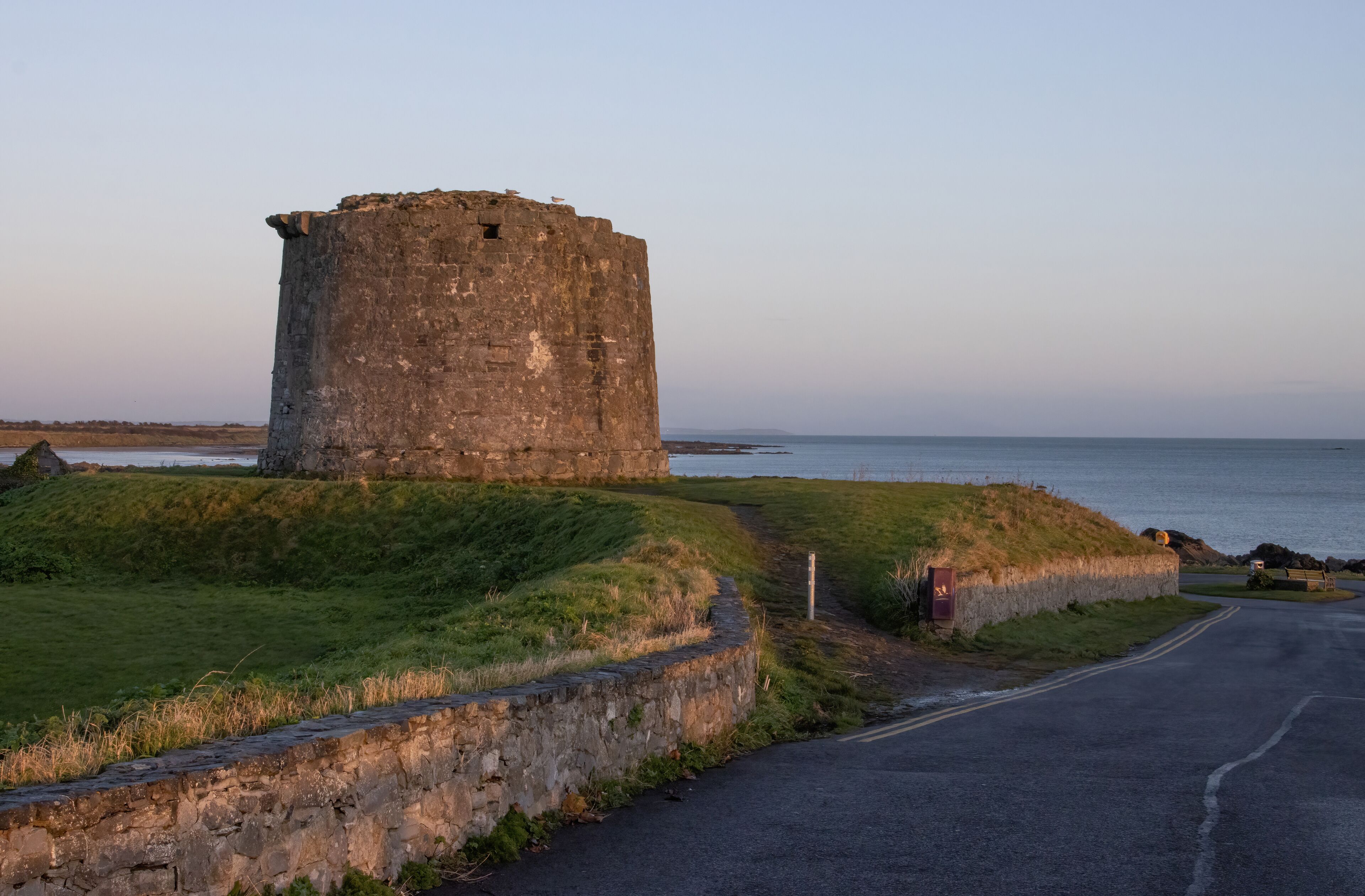 A coastal stone tower with a grassy embankment and a paved road nearby.