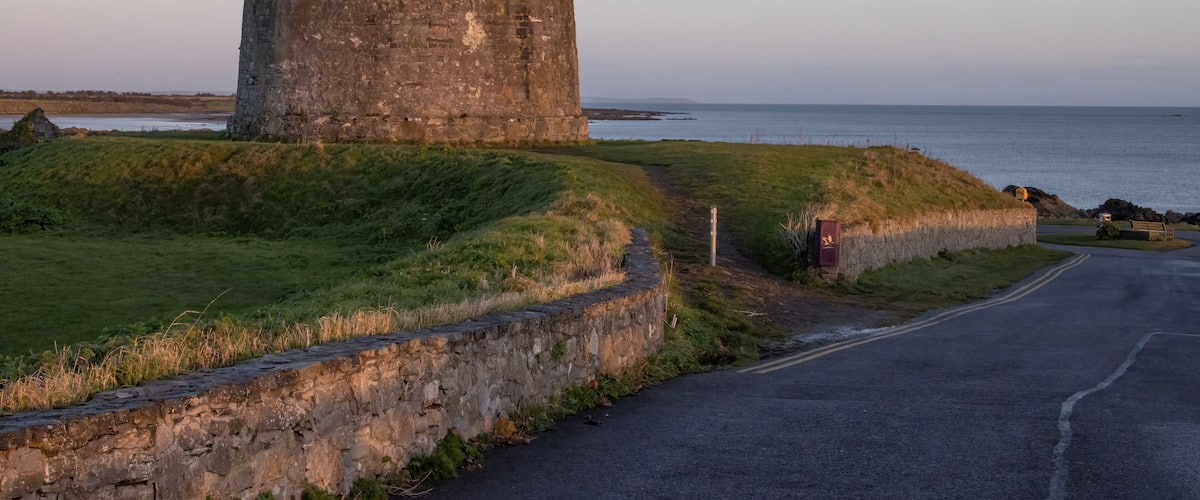 A coastal stone tower with a grassy embankment and a paved road nearby.