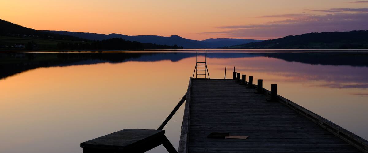 A wooden dock extending into a calm lake at sunset with mountains in the background.
