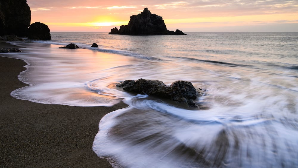 A beach with waves, a sunset, and rocky formations.