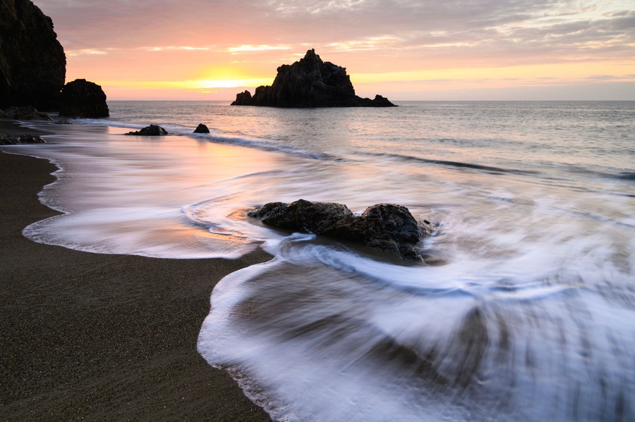 A beach with waves, a sunset, and rocky formations.