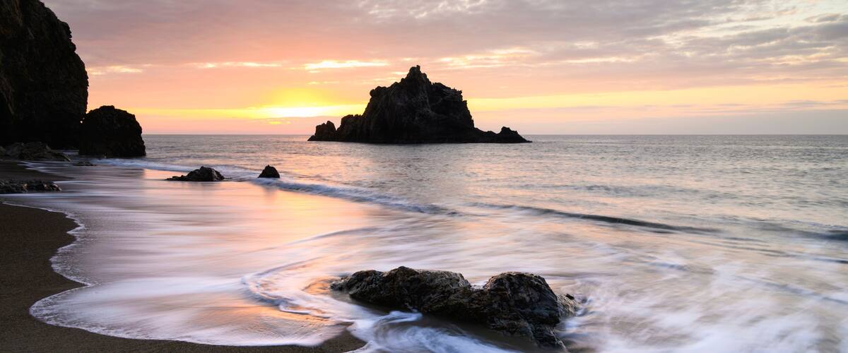 A beach with waves, a sunset, and rocky formations.