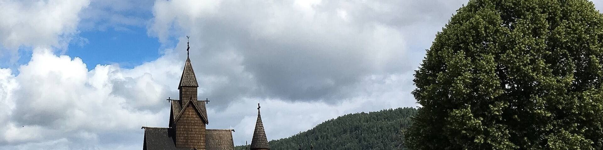 A traditional wooden church with a steep roof and multiple spires, surrounded by lush green fields and a large tree.