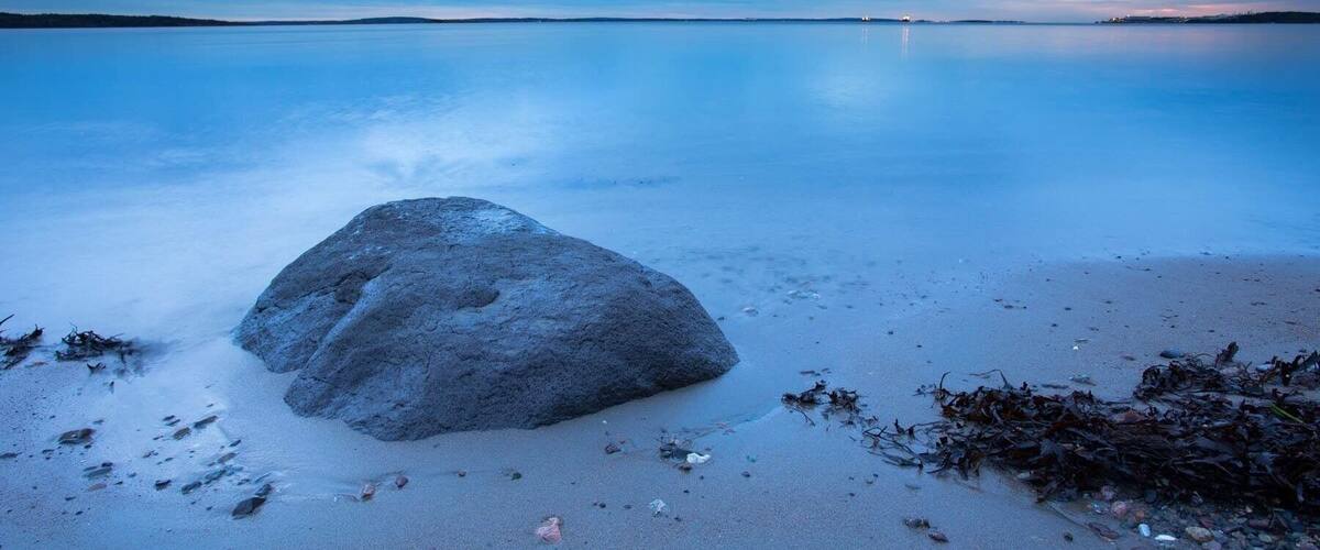 A calm lake with a large rock in the foreground and a clear sky.