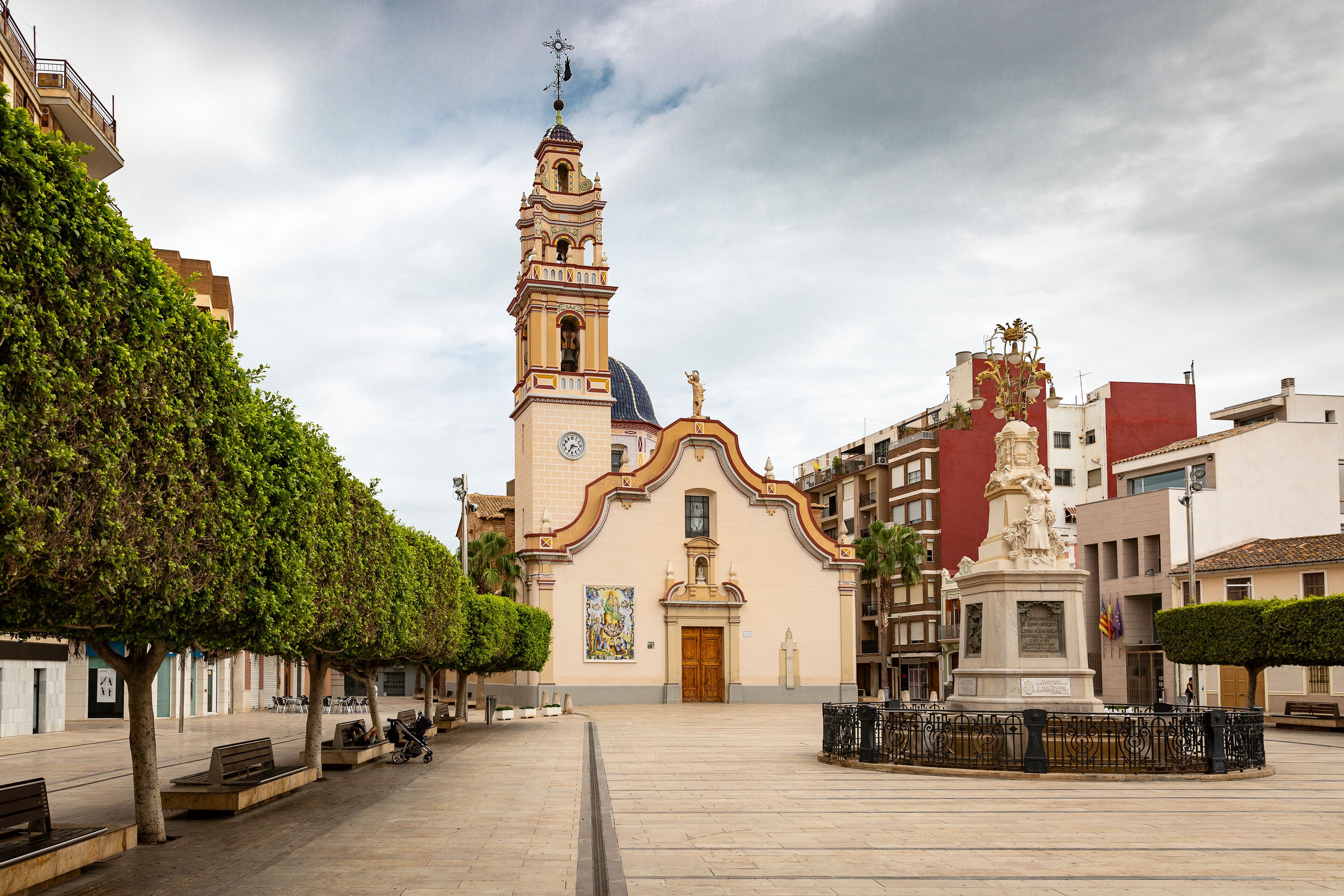 A church with a bell tower, a statue, and a plaza with benches and trees.
