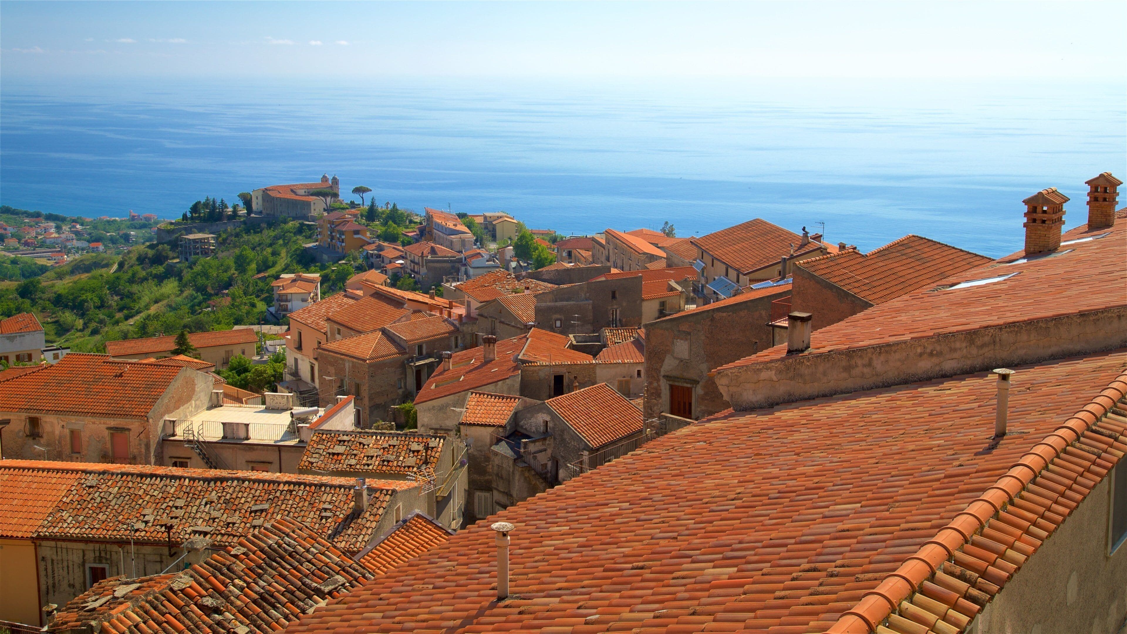 A coastal town with terracotta-roofed buildings, a clear view of the sea, and a distant church.