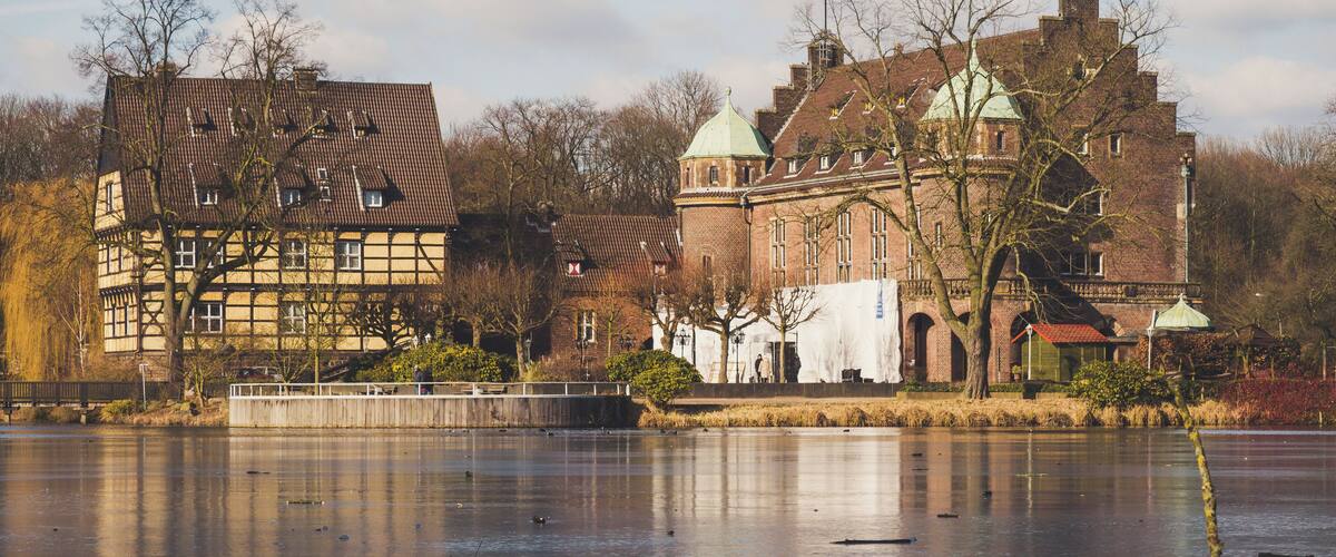 A frozen river with a wooden pier and a large brick building with a green dome in the background.