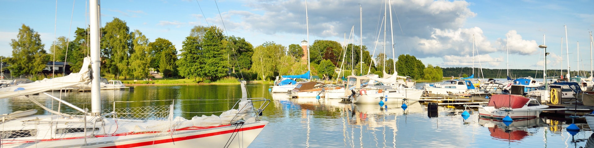 Un puerto deportivo con barcos amarrados a un muelle de madera, rodeado de árboles y un lago tranquilo bajo un cielo parcialmente nublado.