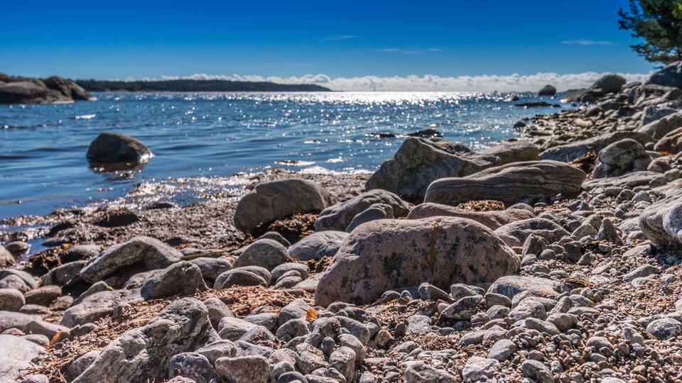 A rocky shoreline with large boulders and pebbles, calm water reflecting the sky, and a distant tree line.