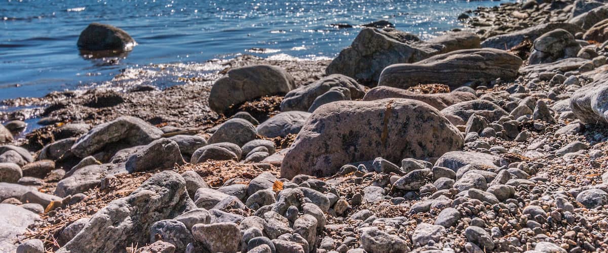 A rocky shoreline with large boulders and pebbles, calm water reflecting the sky, and a distant tree line.