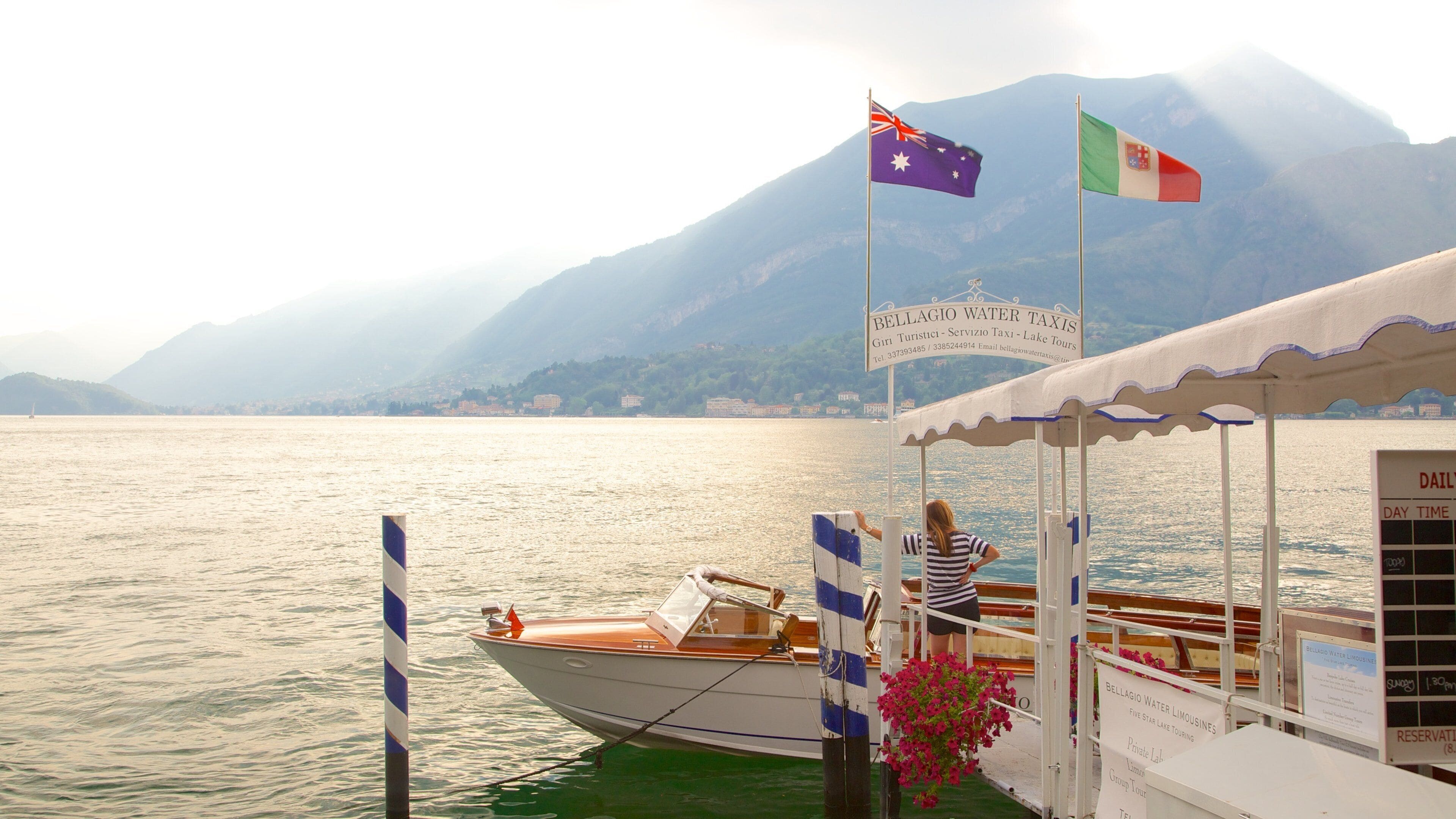 Un barco atracado en un muelle con banderas, un cartel de Bellagio Water Taxis y montañas al fondo.