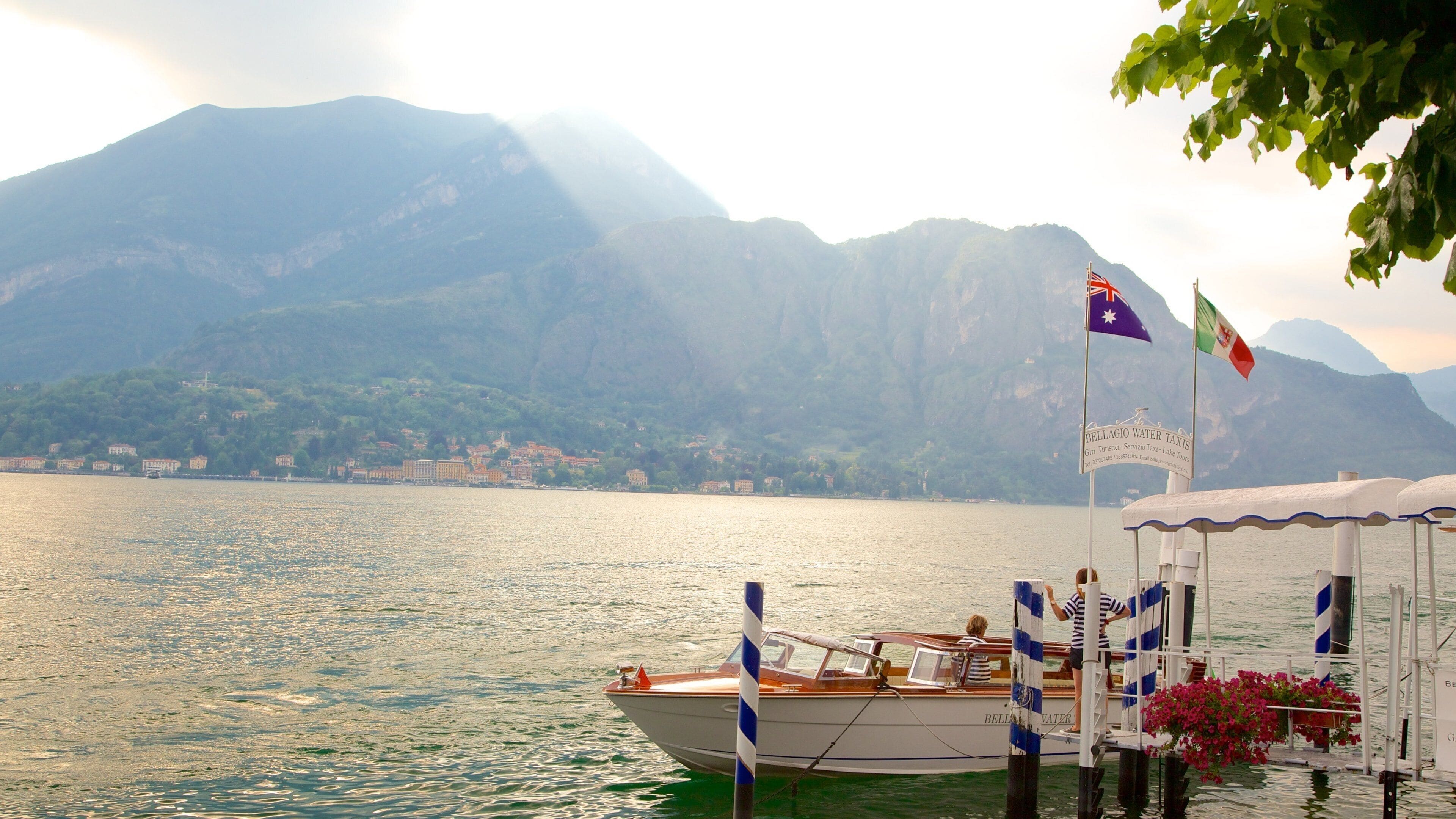 A boat docked at a pier with flags, a mountain range in the background, and a body of water reflecting sunlight.