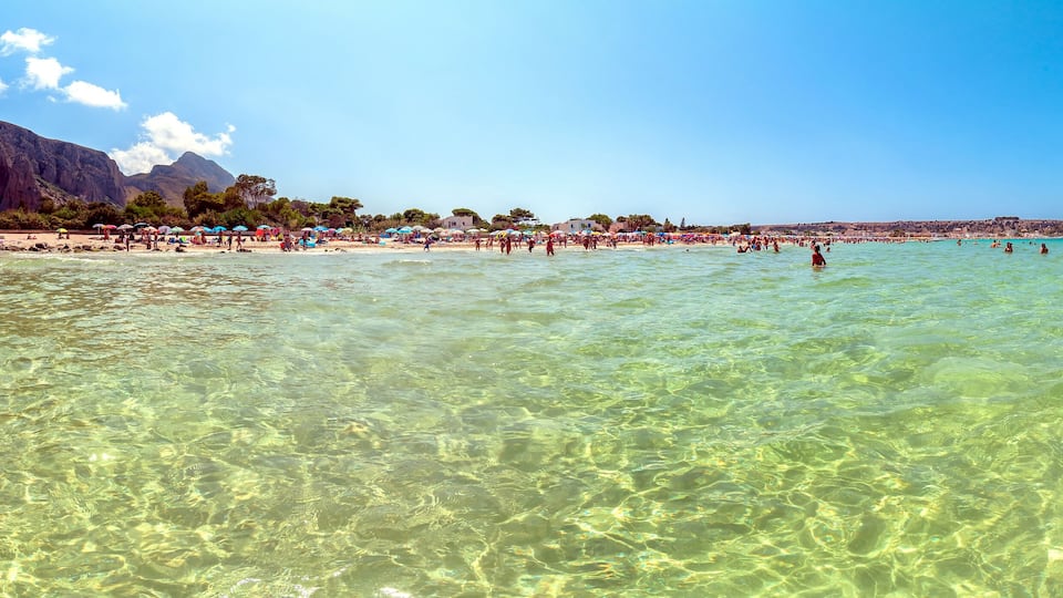 A beach with clear turquoise water, a rocky mountain in the background, and a crowd of people.