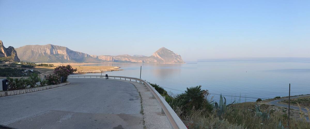 A coastal road with a guardrail, a calm sea, and rocky cliffs.