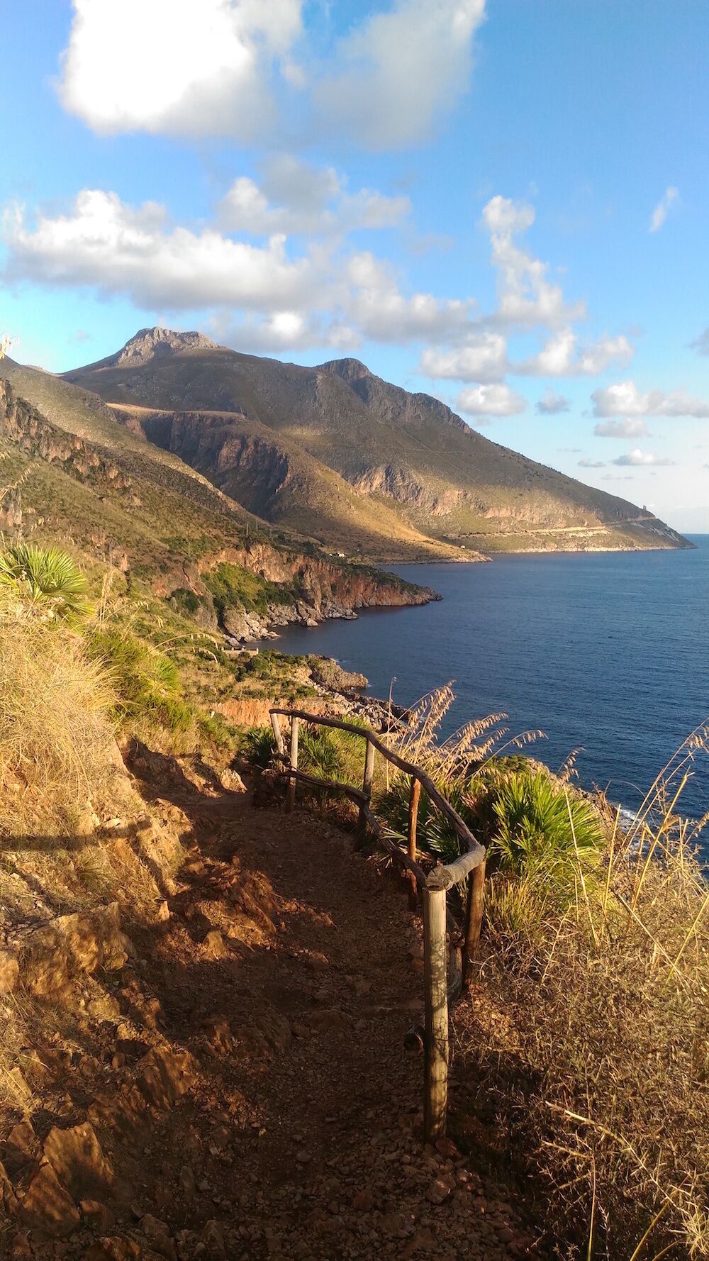 Uma trilha costeira com guarda-corpo de madeira, terreno rochoso e uma montanha ao fundo.