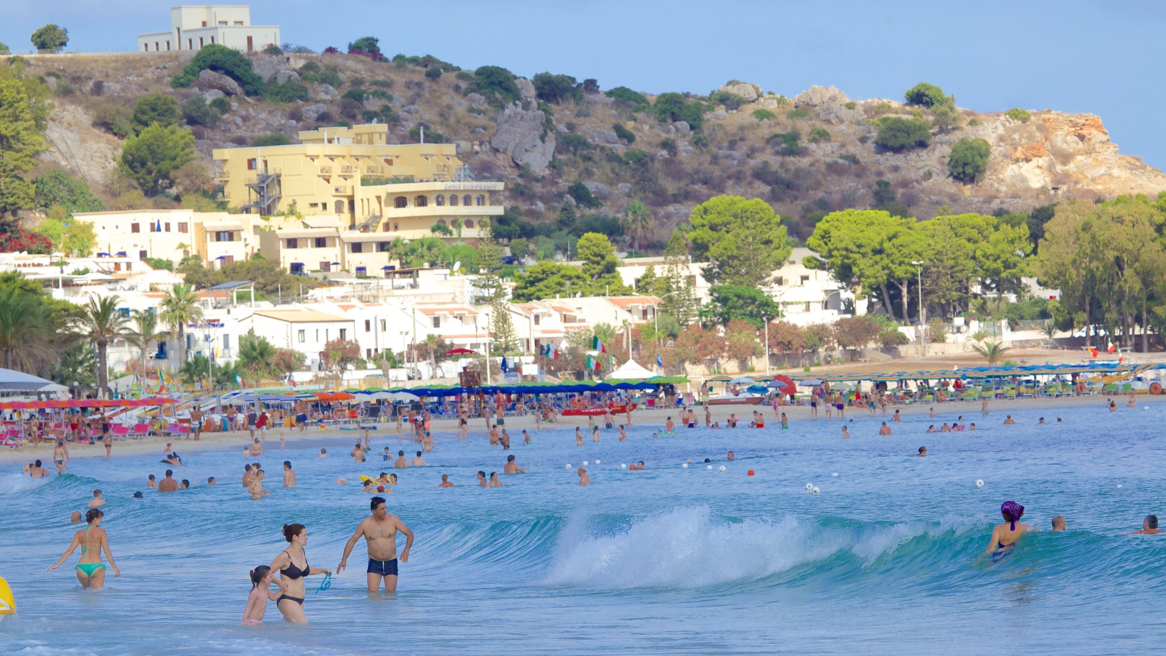 Praia lotada com pessoas nadando e tomando sol, cercada por edifícios e colinas.