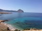 A coastal beach with clear turquoise waters, rocky outcrops, and a distant mountain.