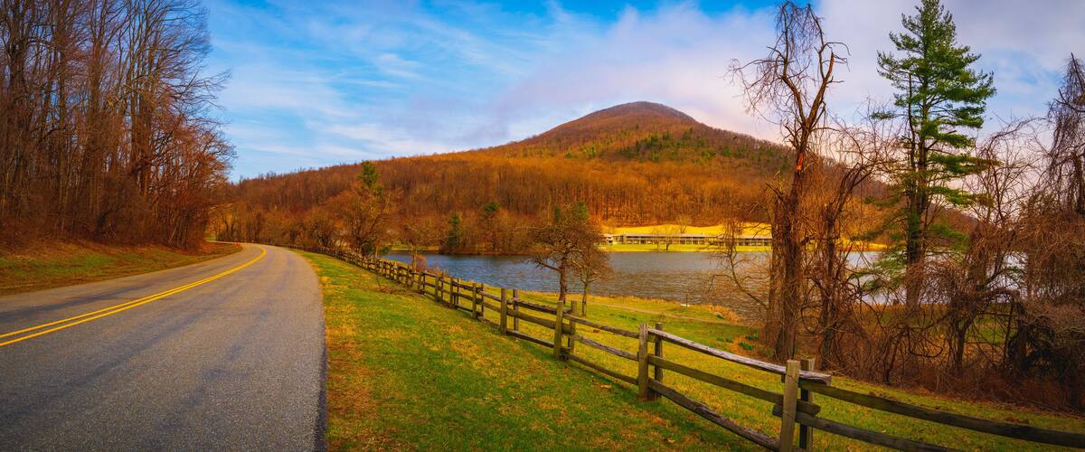A winding road with a wooden fence, a lake, and a forested hill in the background.