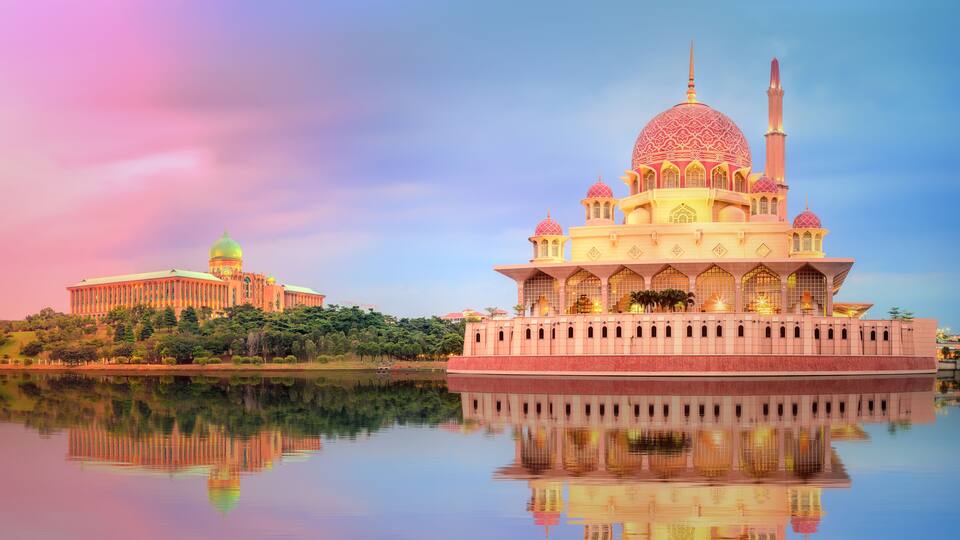 A large mosque with a golden dome and minarets, reflecting in a calm body of water, with a smaller building in the background.