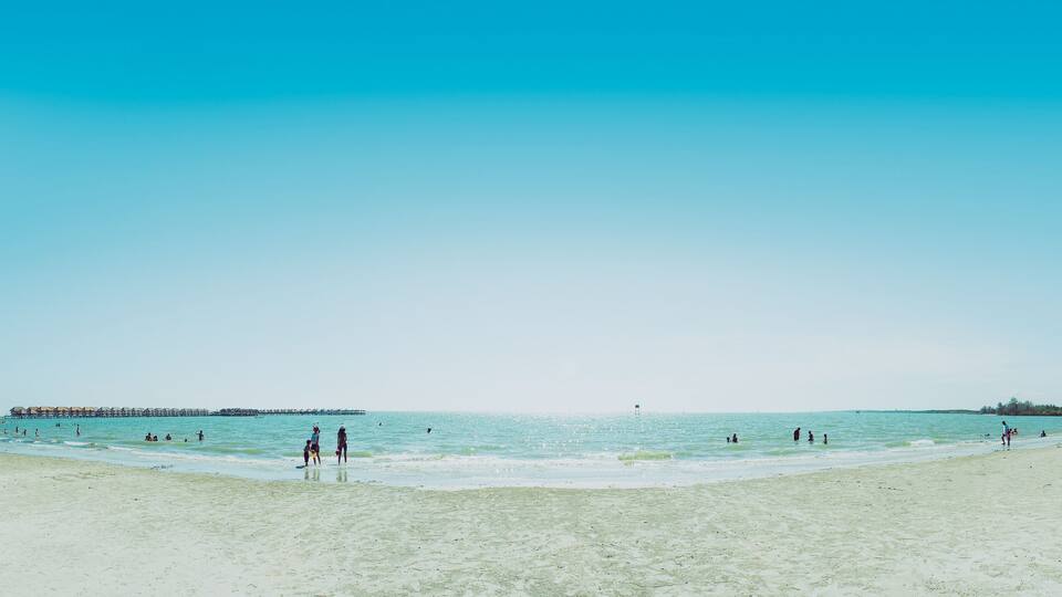 A beach with clear blue water, people swimming and walking on the shore, and a pier in the distance.