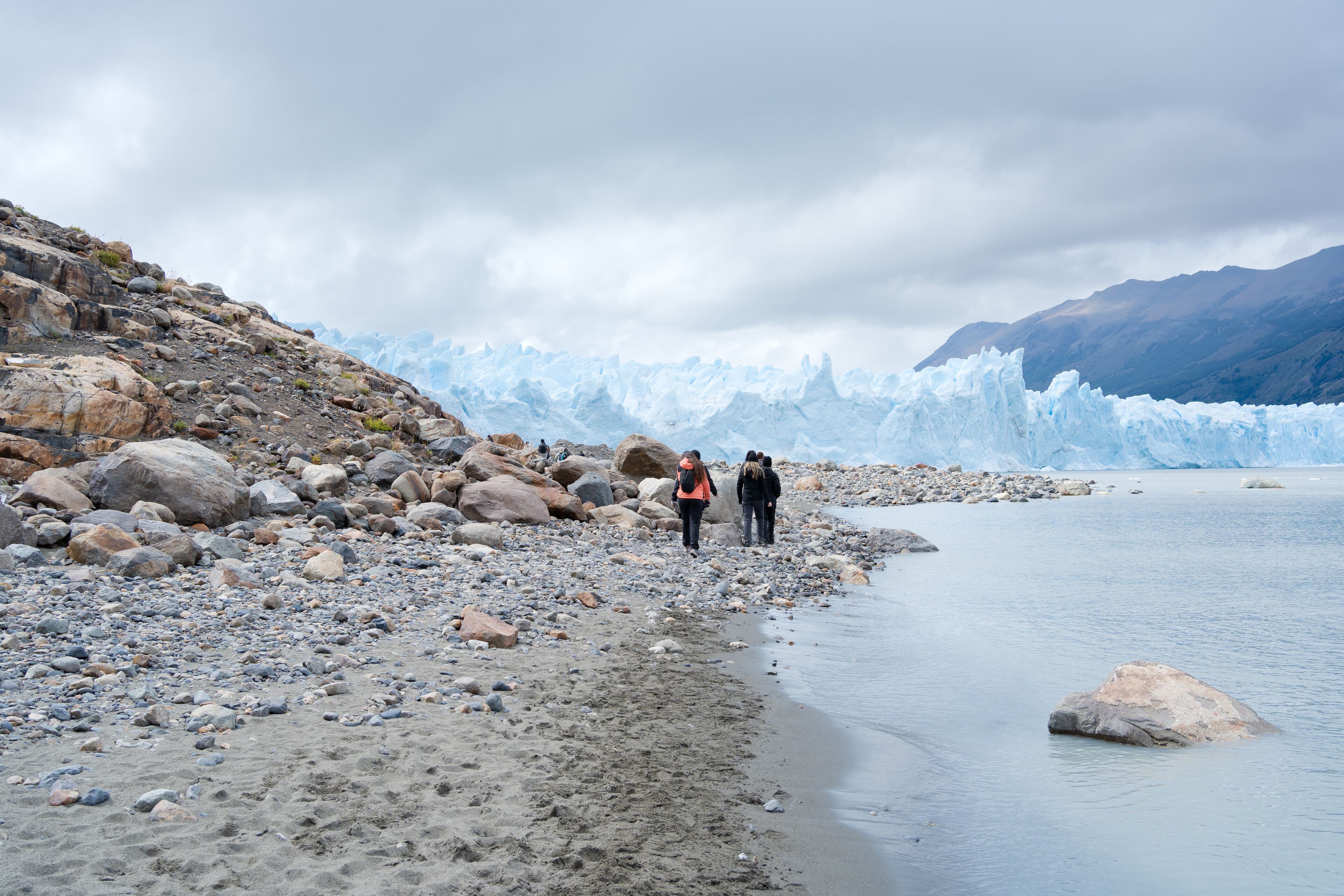 Un grupo de mujeres y hombres se dirigen caminando por la orilla de la playa para llegar y recorrer el imponente glaciar Perito Moreno de Argentina.
