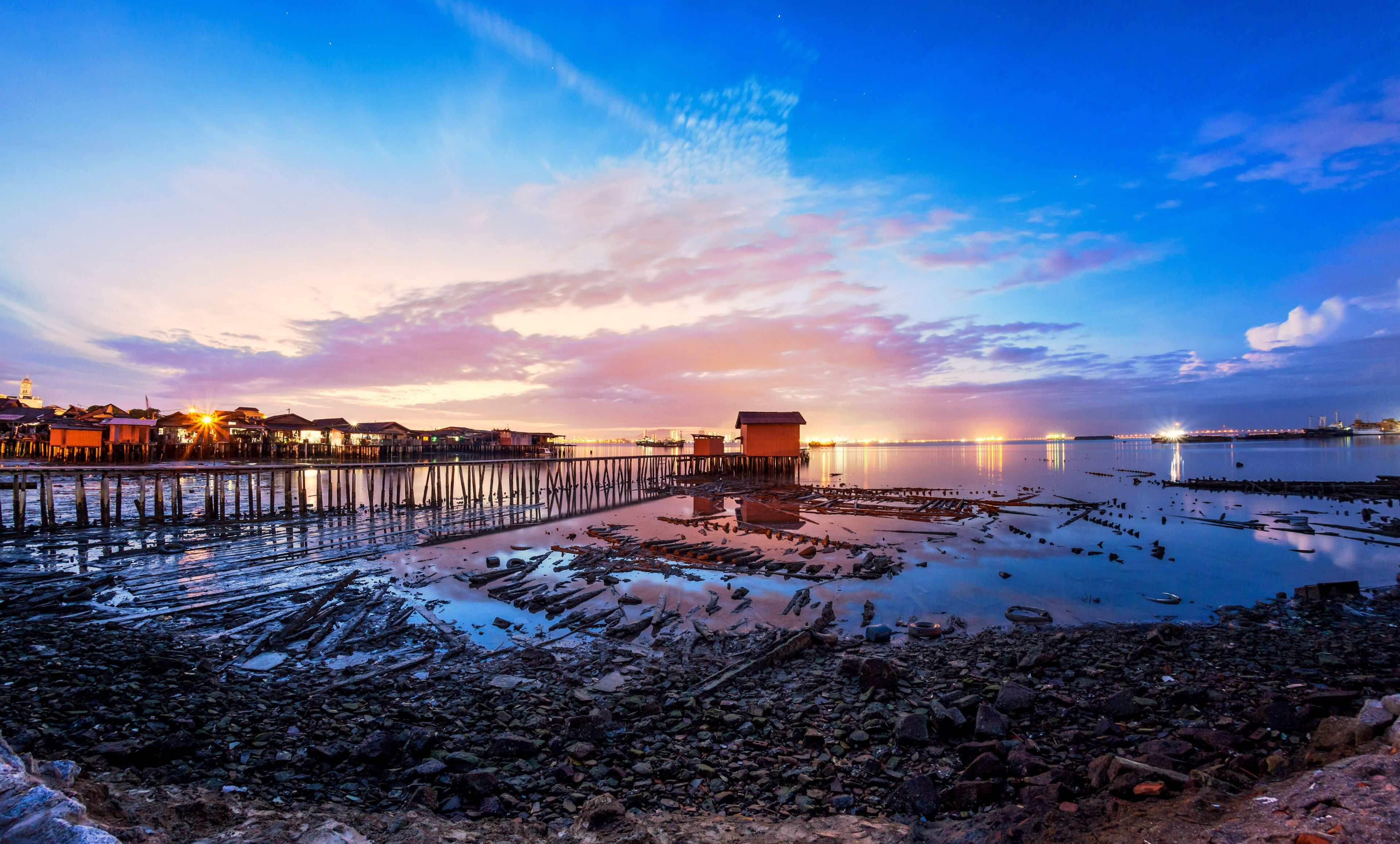 A coastal scene with a pier extending into calm waters, a small wooden structure on the shore, and a distant city skyline at sunset.
