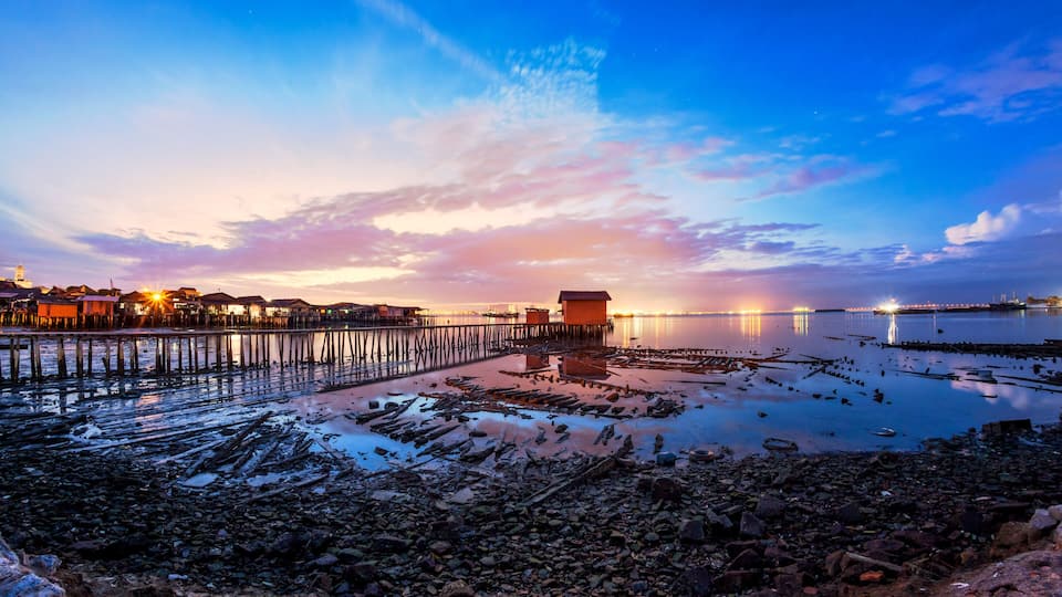 A coastal scene with a pier extending into calm waters, a small wooden structure on the shore, and a distant city skyline at sunset.