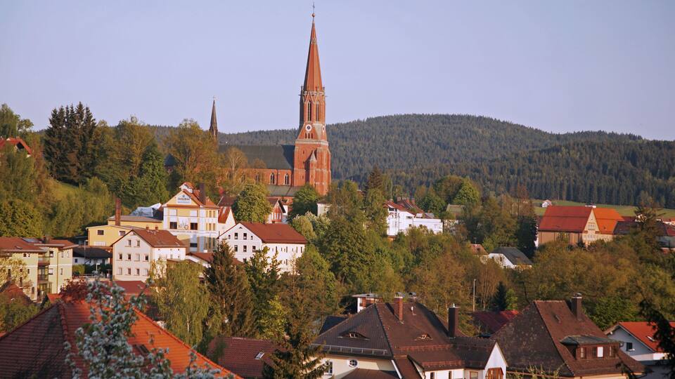 A town with a prominent church tower, surrounded by residential buildings and a forested hill in the background.