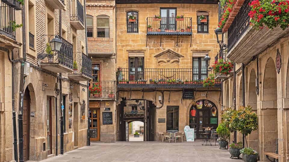 A narrow street with historic buildings, balconies with flower boxes, and outdoor seating areas.