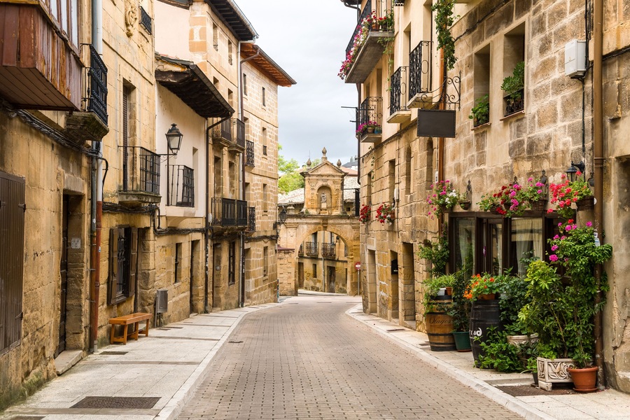 Una estrecha calle empedrada flanqueada por edificios históricos de piedra, algunos con balcones y jardineras.