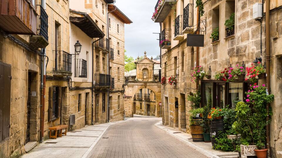 A narrow cobblestone street lined with historic stone buildings, some with balconies and flower boxes.