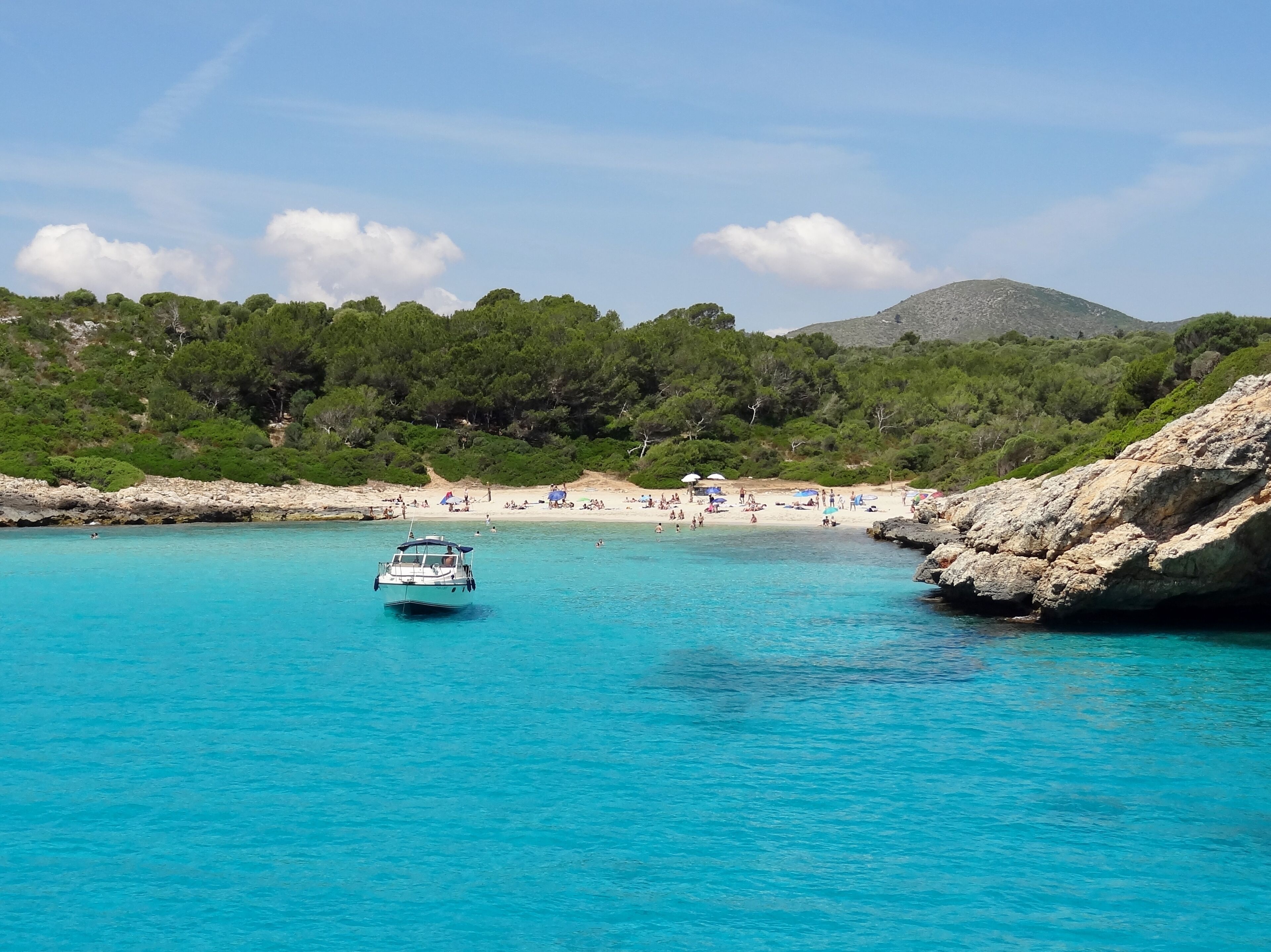 A boat on turquoise waters, a sandy beach, and a rocky coastline with trees.