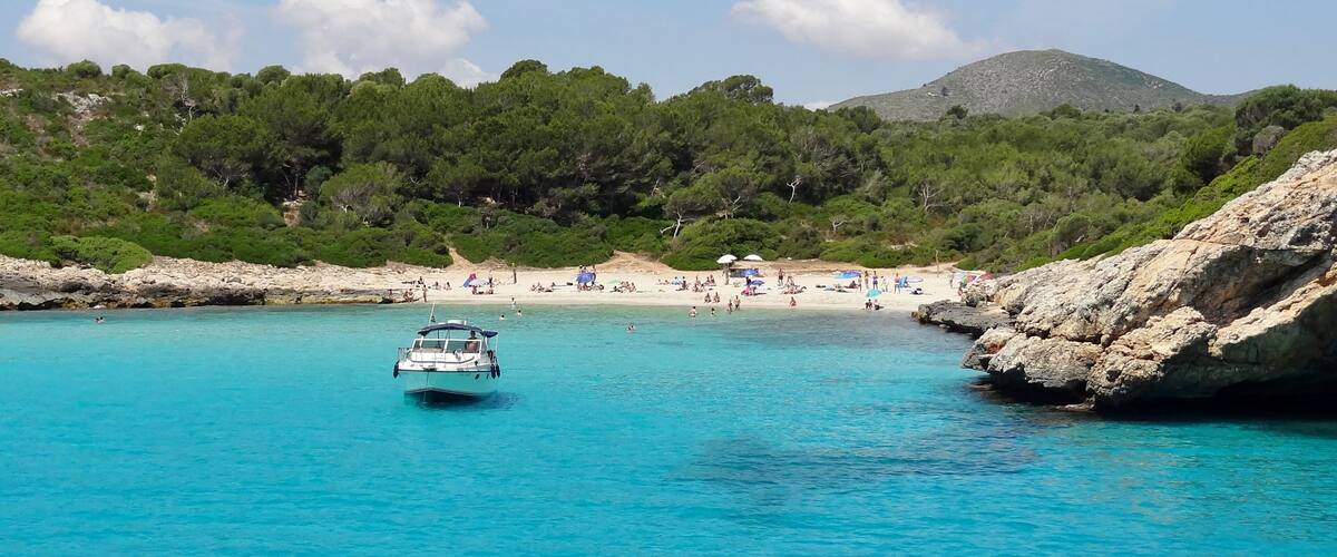 Un bote sobre aguas turquesas, una playa de arena y un litoral rocoso con árboles.