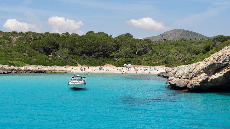 A boat on turquoise waters, a sandy beach, and a rocky coastline with trees.