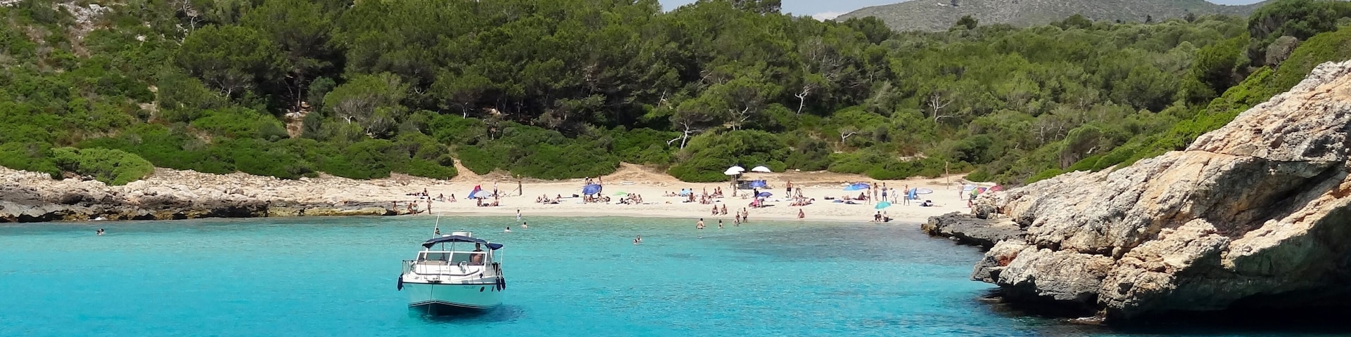 Un bote sobre aguas turquesas, una playa de arena y un litoral rocoso con árboles.