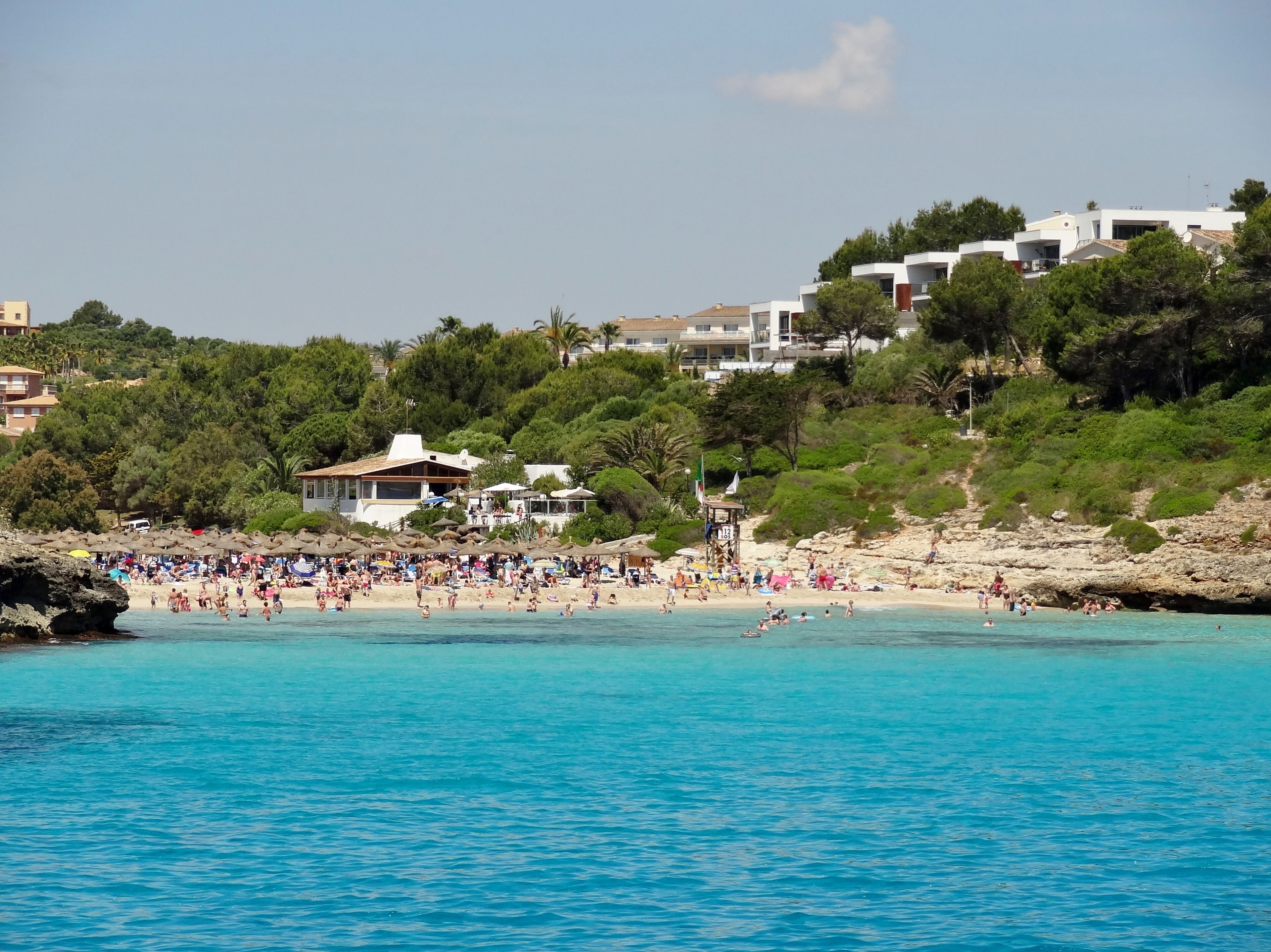 A crowded beach with clear turquoise water, surrounded by lush greenery and modern buildings.