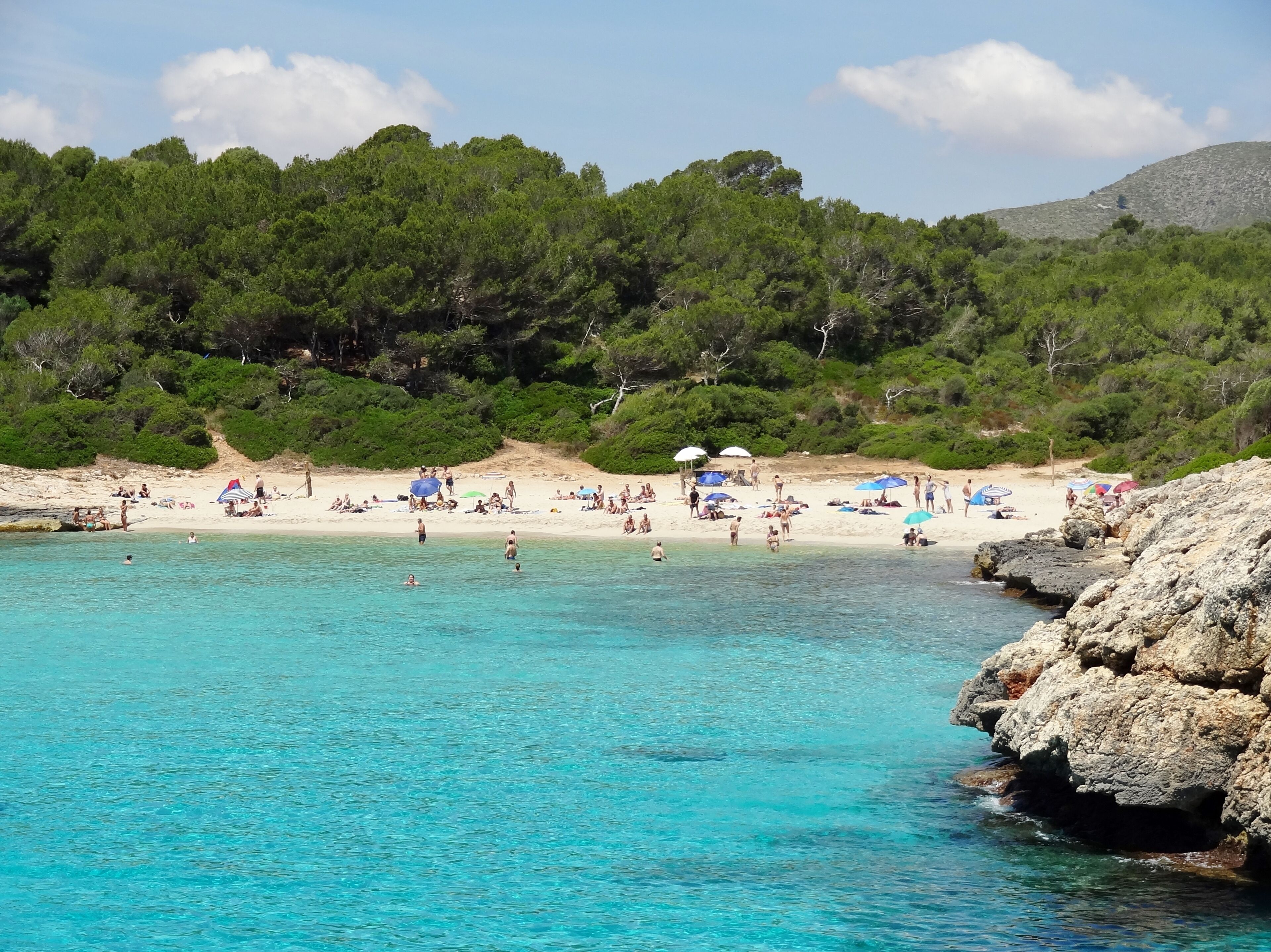 Una playa con aguas turquesas cristalinas, gente tomando el sol y colinas verdes exuberantes al fondo.