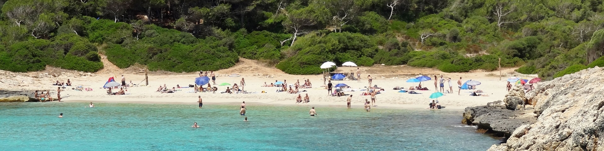 Una playa con aguas turquesas cristalinas, gente tomando el sol y colinas verdes exuberantes al fondo.