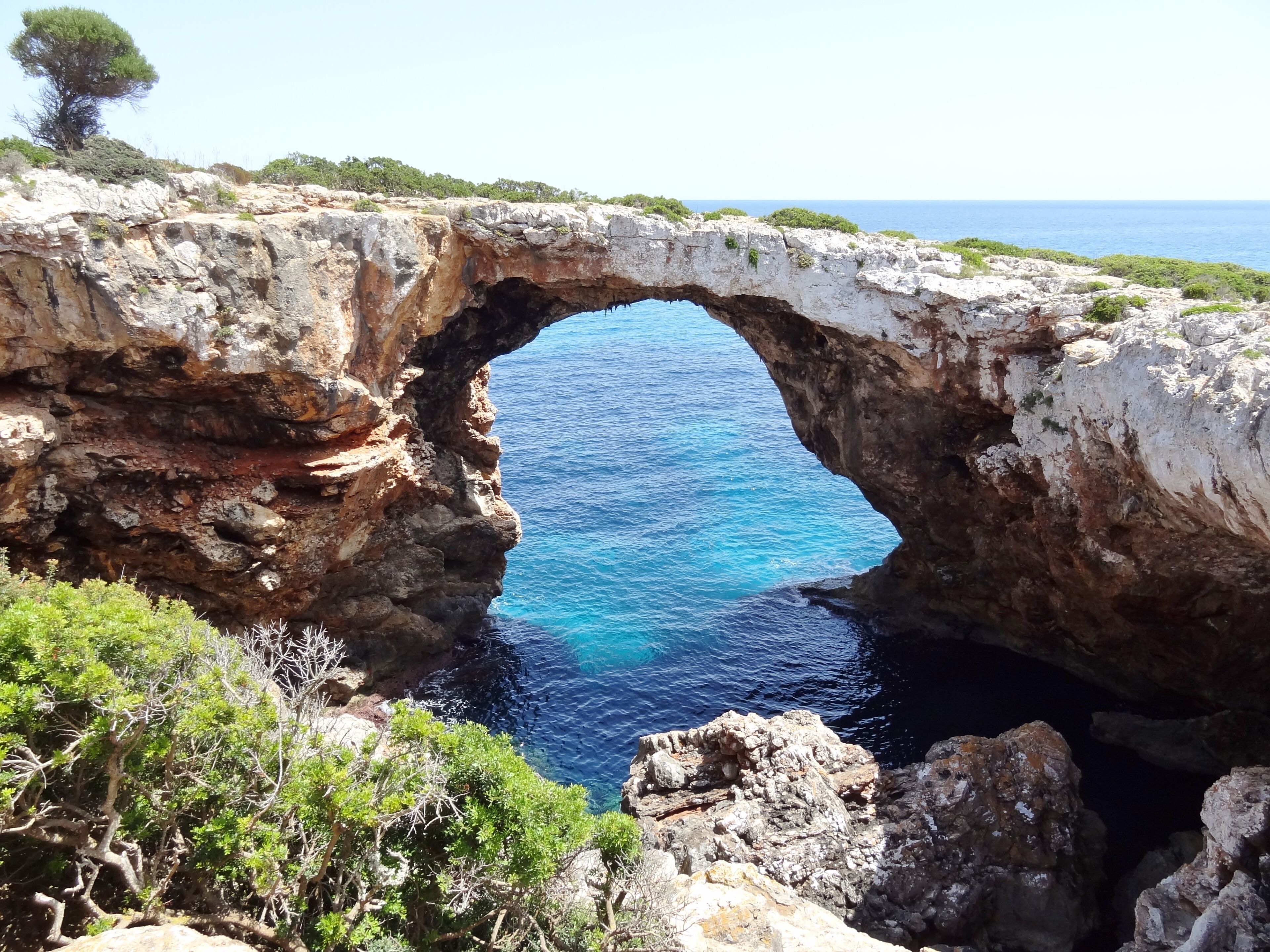 A natural rock arch over a clear blue sea, surrounded by rocky terrain and sparse vegetation.