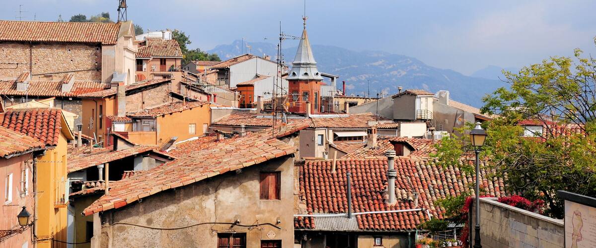 A historic town with terracotta-roofed buildings, a bell tower, and mountains in the background.