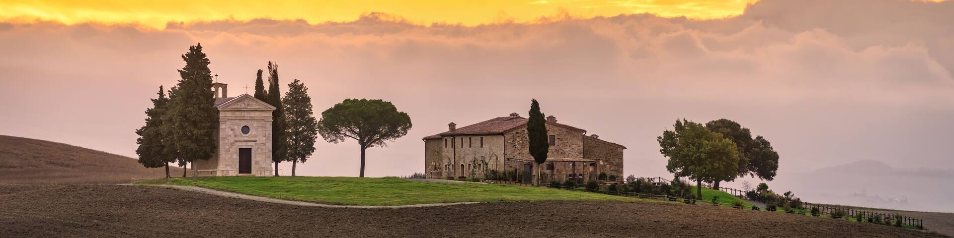 A rural landscape with a church, a stone building, and trees under a sunset sky.
