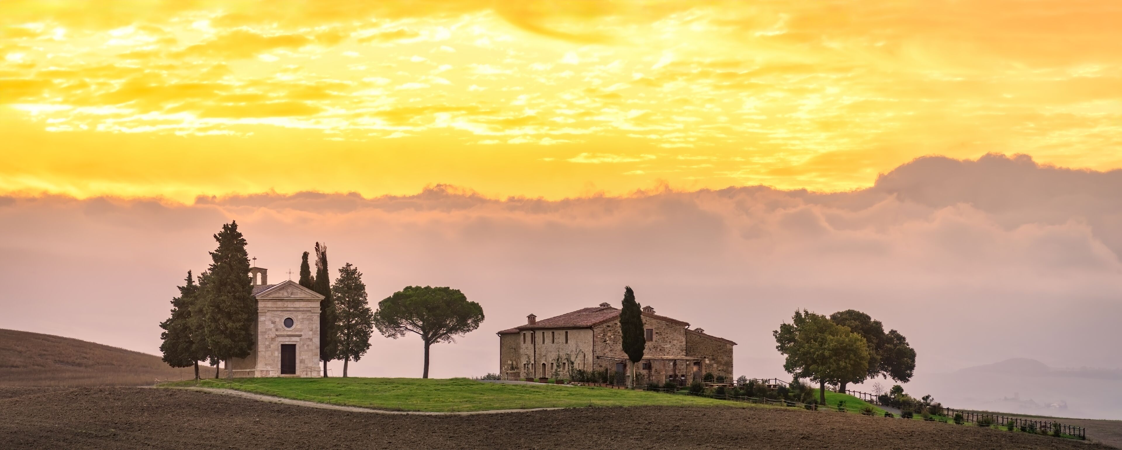 A rural landscape with a church, a stone building, and trees under a sunset sky.
