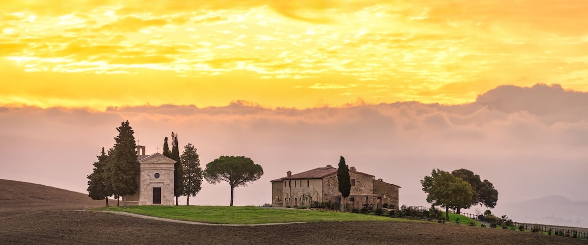 A rural landscape with a church, a stone building, and trees under a sunset sky.