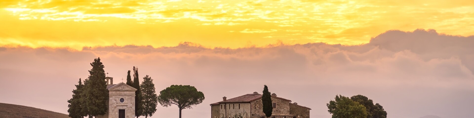 A rural landscape with a church, a stone building, and trees under a sunset sky.
