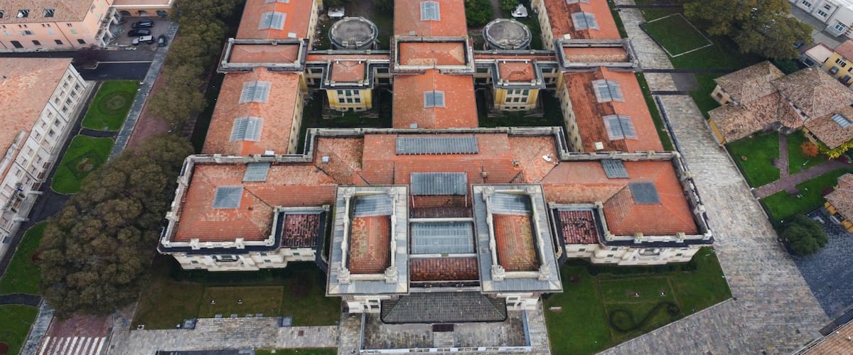 An aerial view of a historic walled city with a central courtyard, surrounded by buildings with red-tiled roofs.