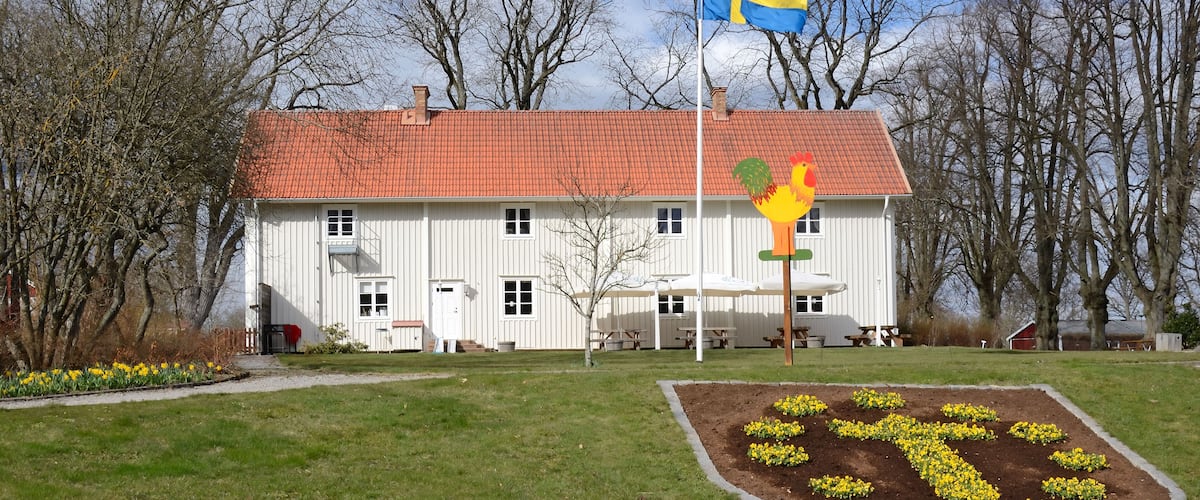 A white building with a red roof, a Swedish flag, and a rooster sign.