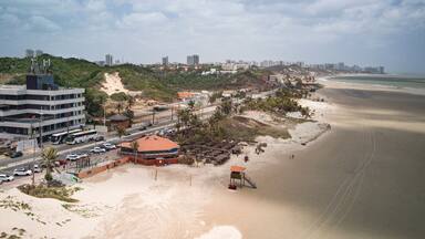 Aerial view of Calhau Beach in São Luís, Maranhão, Brazil, showing palm trees, sandy shore, beach umbrellas, and urban landscape along the coastline on a cloudy day